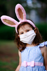 Little toddler girl with bunny ears and surgical face mask hunting for Easter eggs during coronavirus quarantine - 337056998