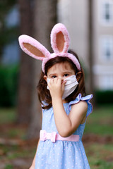 Little toddler girl with bunny ears and surgical face mask hunting for Easter eggs during coronavirus quarantine - 337057100