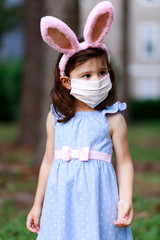 Little toddler girl with bunny ears and surgical face mask hunting for Easter eggs during coronavirus quarantine - 337057137