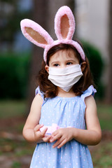 Little toddler girl with bunny ears and surgical face mask hunting for Easter eggs during coronavirus quarantine - 337057196