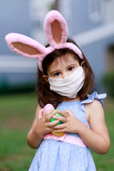 Little toddler girl with bunny ears and surgical face mask hunting for Easter eggs during coronavirus quarantine - 337057372