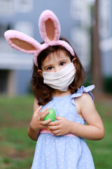 Little toddler girl with bunny ears and surgical face mask hunting for Easter eggs during coronavirus quarantine - 337057376