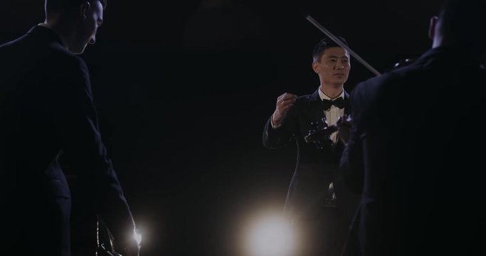 Rear of musicians playing on violins. Back view on men and woman performing music. Joyful happy smiled Asian young male conductor controlling the classical symphony orchesra at concert.