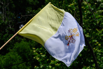 Vatican national flag blowing in the wind in direct sunlight towards blurred green trees and leaves
