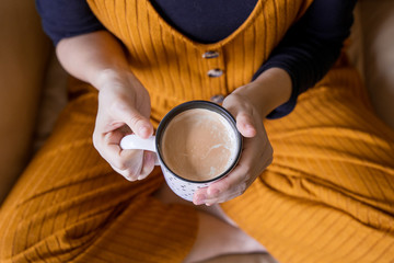Obraz na płótnie Canvas Top view of unrecognizable woman holding a hot cup of coffee at home. Detail of warm cup of latte on white mug. Healthy lifestyle. Woman drinking exotic coffee for balance diet. Beverage concept.