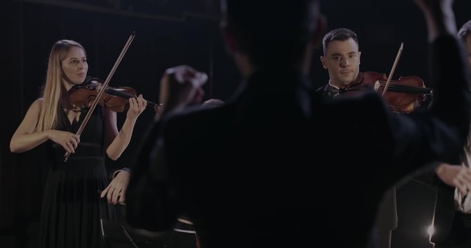 Back view of male conductor controlling band. Quartet of classical male and female musicians performing music on dark stage in spotlight. Orchestra playing on violins and cello. Rear of man directing.