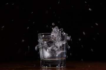 A glass of water on a black background, into which pieces of ice are thrown and splashes fly