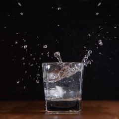 A glass of water on a black background, into which pieces of ice are thrown and splashes fly