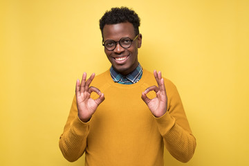 Portrait of happy african american man in glasses showing ok sign and smiling, over yellow...