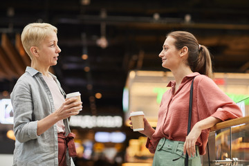 Two women talking to each other and drinking coffee while standing in the mall