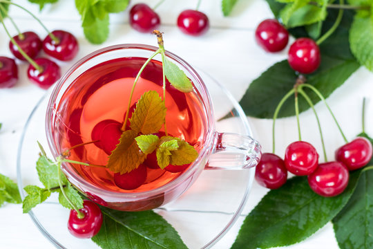 Fresh fruit cherry drink in transparent glass cup surrounded by cherries on the white wooden background