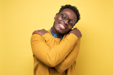 Pleased african american man in yellow clothes and glasses hugs himself, has high self esteem....