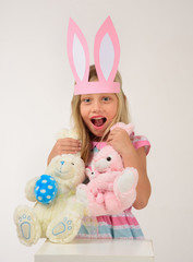 Beautiful girl with pink paper bunny ears holds 2 rabbits by the ears with a look of astonishment, on a gray background.