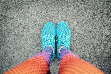 Selfie of female legs in blue sneakers and multi-colored leggings on the pavement, top view