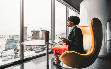 A ravishing African-American businesswoman on a yellow armchair is using her tablet PC; biracial woman entrepreneur using a digital tablet being on the top floor of a luxury business office skyscraper