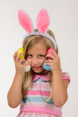 Pretty girl with pink bunny ears plays with 4 little colorful eggs holding it against her face, on a gray background, the girl smiles.