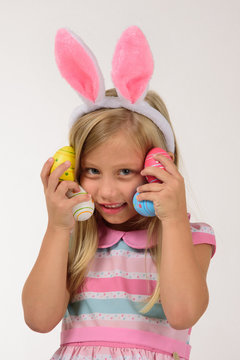 Pretty girl with pink bunny ears plays with 4 little colorful eggs holding it against her face, on a gray background, the girl smiles.