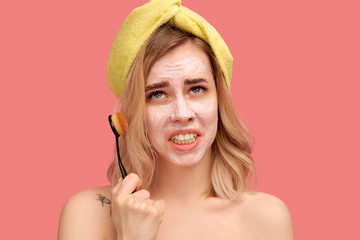 Unhappy young girl holds brush to clean face, has dissatisfied expression , asks God what this punishment is for.. Beautiful woman with yellow hat or towel stands naked against pink wall Studio