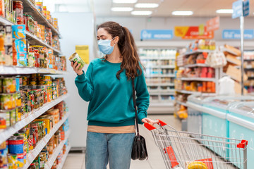 Shopping. A young pretty Caucasian woman in a medical mask buys a can of canned peas at a grocery store. The concept of buying products and the coronovirus pandemic