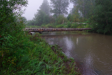 Side view of a summer landscape with river and small old wooden suspension bridge for people traveling across the river