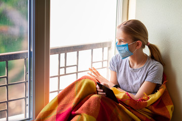 Sad little girl in a mask sitting on the window with e-book reading at home isolated. Coronavirus quarantine concept.