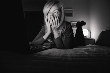 Blonde girl is lying on the bed in the dark and looking at the laptop screen, she is scared and covers her face with her hands. Black and white portrait.