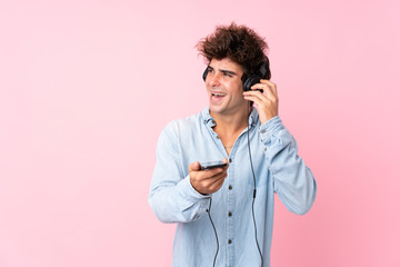 Young caucasian man over isolated pink background using the mobile with headphones and singing