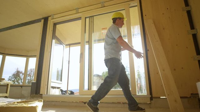 SLOW MOTION, LOW ANGLE, SUN FLARE: Caucasian contractor picks up a plank and carries it across the ground floor of a contemporary CLT house. Golden sun rays shine on builder working in timber house.