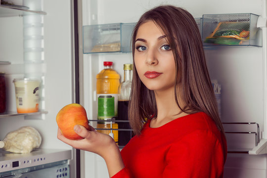 Healthy Eating Concept. Diet. Beautiful Young Woman in red dress near the Refrigerator with orange fruit in hand looking at you camera asking her self what to eat.