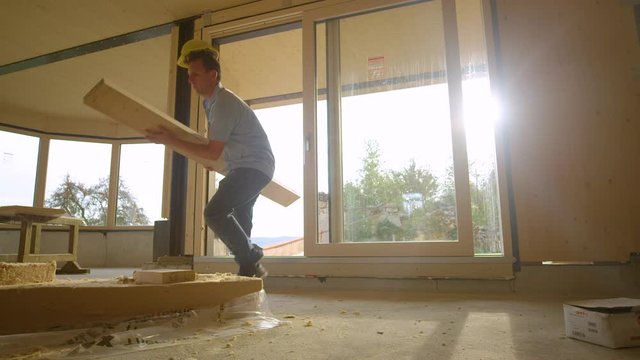 SLOW MOTION LENS FLARE, LOW ANGLE: Young Caucasian man wearing a yellow hard hat and carrying a long plank trips while entering the glued-laminated timber house under construction. Clumsy builder.