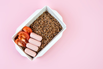 Delivery of a box of cooked buckwheat, sausage and tomato on a pink trend background