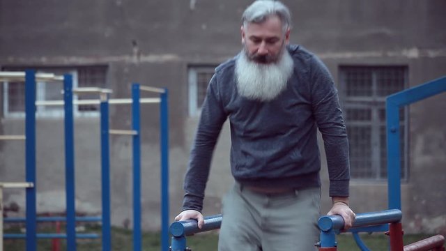 Senior bearded man with strong muscular arms exercising outdoors, doing pull ups, an elderly male performing pulling exercise on horizontal bar