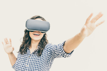 Positive female user in VR goggles touching air and smiling. Young woman in virtual reality headset standing isolated over white background. Augmented reality concept