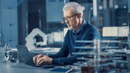 Professional Electronics Design Engineer Wearing Glasses Works on Computer in Research Facility. In the Background Motherboards, Circuit Boards and Computer Components in the High Tech Facility.