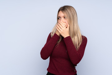 Young blonde woman over isolated blue background covering mouth and looking to the side