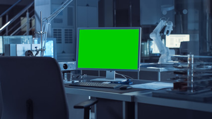  On the Desk Computer with Isolated Green Mock-up Screen Display. In the Background Robot Arm Concept Standing in Heavy the Dark.Industry Engineering Facility