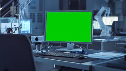 Fototapeta premium On the Desk Computer with Isolated Green Mock-up Screen Display. In the Background Robot Arm Concept Standing in Heavy the Dark.Industry Engineering Facility