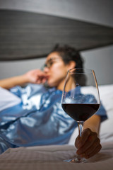 A distraught woman wearing satin pajamas sits in bed with a glass of wine