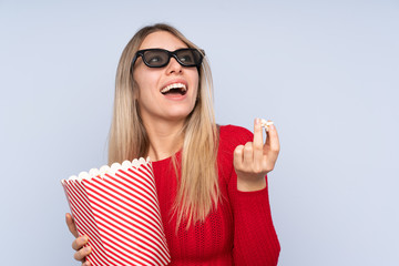 Young blonde woman over isolated blue background with 3d glasses and holding a big bucket of popcorns