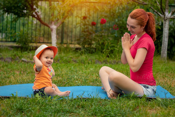 Young woman doing yoga with baby in the summer park. Family outdoors. Parent with child spend time together.