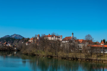 Obraz premium Die Altstadt von Füssen auf der anderen Seite des Lechs mit Hohem Schloss, St. Mang mit Kloster und Kirche und der Spitalkirche bei blauem Himmel