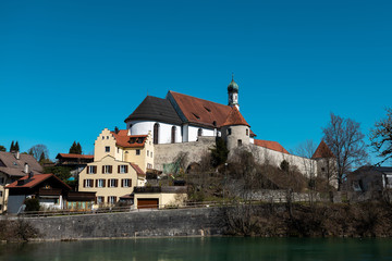 Fototapeta premium Franziskanerklosterkirche in Füssen mit Teilen der alten Stadtmauer und dem Lech im Vordergrund im Frühjahr bei blauem Himmel
