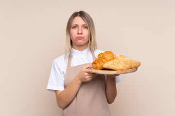 Young blonde girl with apron. Female baker holding a table with several breads with sad expression