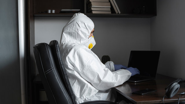 Man in protective suit, medical mask and rubber gloves sits at home and works with laptop at the table during quarantine. Man, designer, artist, architect, businessman at remote work in a pandemic