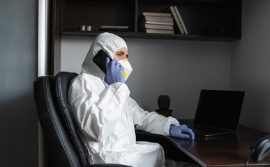 Fototapeta na wymiar Man in protective suit, medical mask and rubber gloves sits at home and speaking on a phone with friends or business partner during quarantine. Man at remote work in a pandemic covid. Coronavirus.