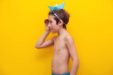 A cheerful boy in swimming trunks and with a paper boat on his head laughs. Travel background with a boy of 9 years. Family travel at sea