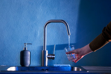 The hand of a young woman pours clean water from under a kitchen faucet into a glass cup on a background of blue wall with empty place for text. Сlean ecology concept in the city