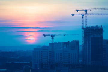 Tower cranes, the construction of a new tall apartment building at a construction site in the city at sunset or sunrise. Profitable investments and renovation program, development, construction