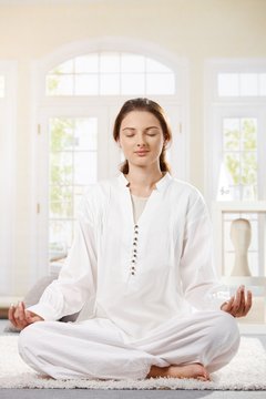 Young woman in white clothes sitting in lotus position, doing yoga, eyes closed, smiling.