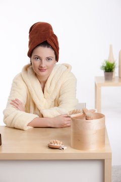 Young attractive teenager sitting at cosmetics desk in bathrobe, hair wrapped in towel, looking at camera, smiling. Isolated on white.